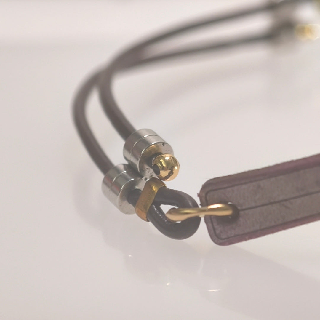 A close-up of a dark brown leather strap accessory with silver and gold metal hardware, resting on a reflective white surface.