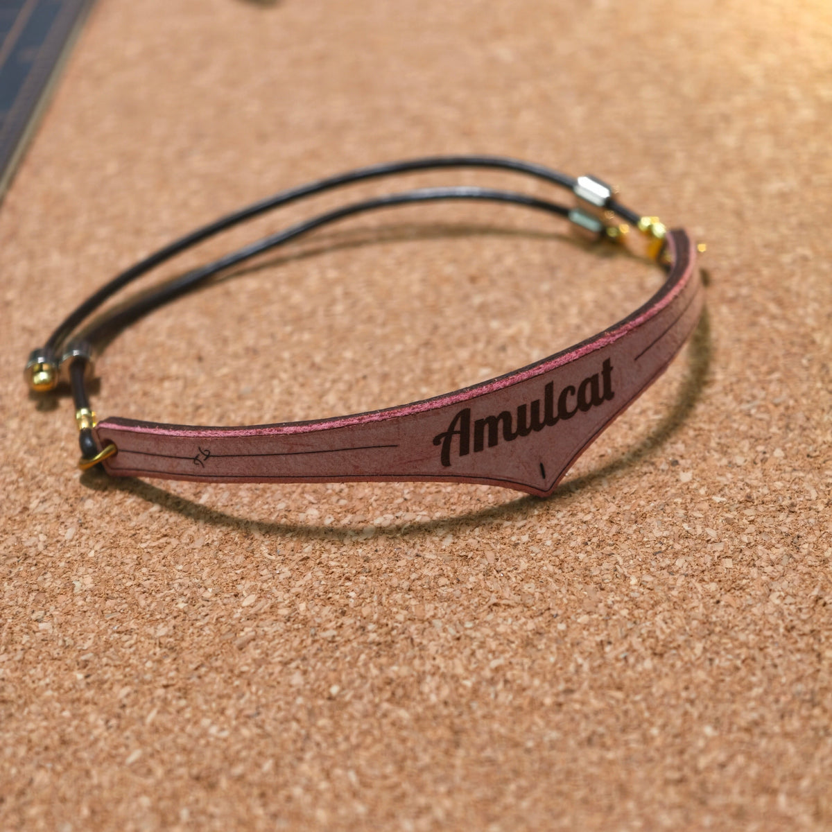 A slim black double-strand cord cat necklet with a central reddish-brown leather tab engraved with the name 'Amulcat', resting on a cork surface.